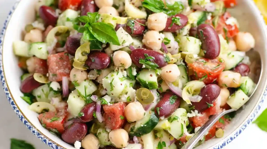 Mediterranean bean salad featuring chickpeas, kidney beans, white cannellini beans, cherry tomatoes, cucumber, bell peppers, kalamata olives, feta cheese, and fresh herbs with lemon vinaigrette in large white bowl.