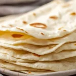 Soft easy flour tortillas from scratch stacked on a cloth with flour and rolling pin nearby